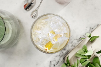A glass with cubed ice and lemon slices placed on a white marble countertop, surrounded by a glass container and greenery.